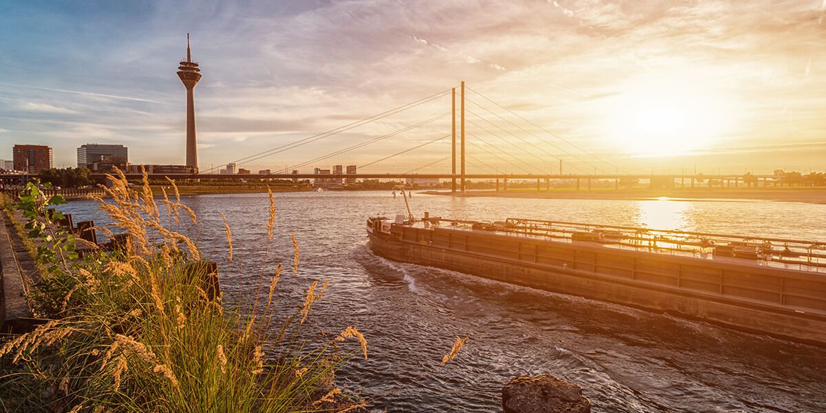 Entdecken Sie ein Schiff auf dem Rhein, welches in Richtung des Fernsehturms fährt bei Sonnenuntergang in Düsseldorf