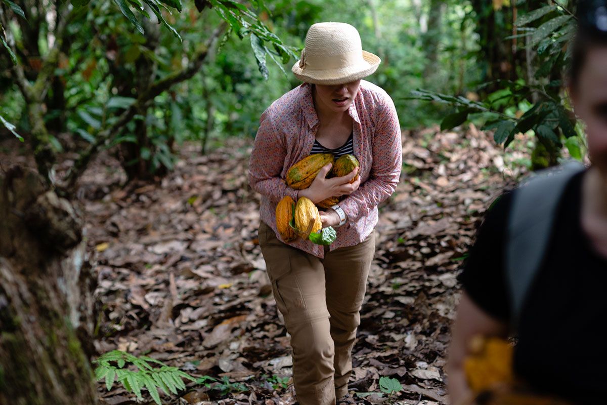 A woman carries several fruits through a forest.